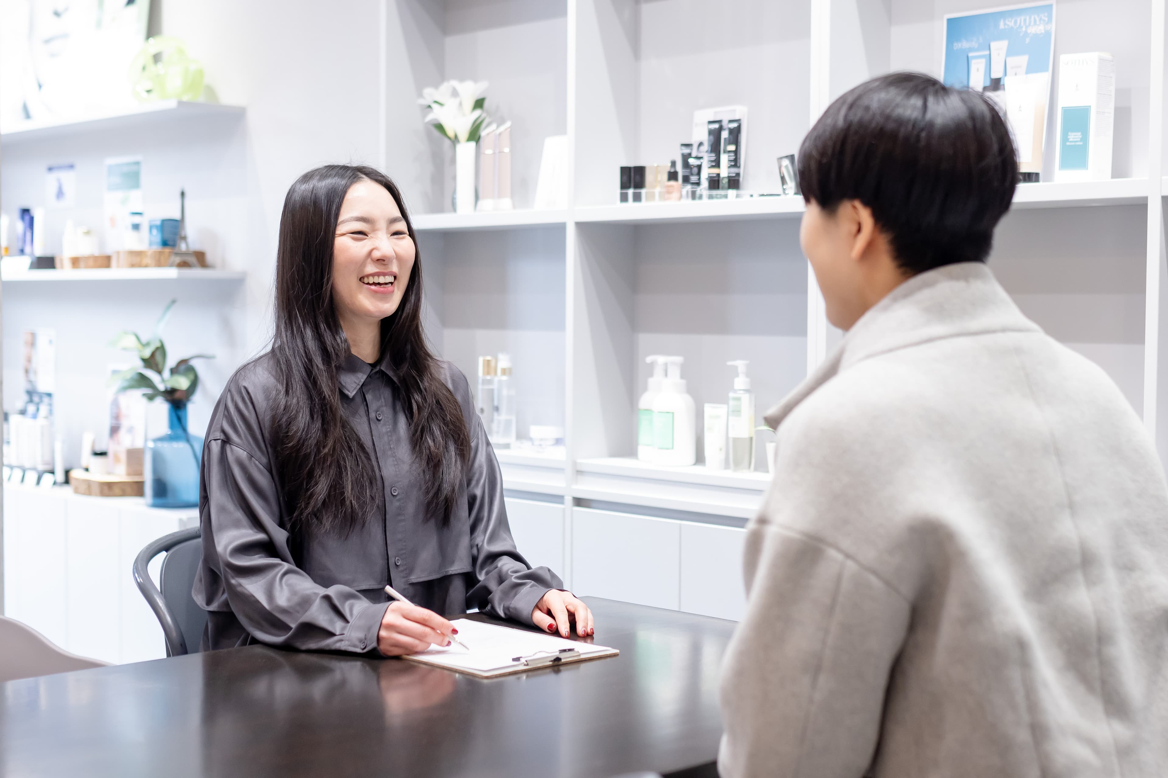 化粧品専門店の知識を活かして肌状態を見極める肌診断サポート - 三田市エステサロン ルナクレッシェンテのスタッフが化粧品棚を背景にお客様の肌質や悩みに合わせた施術とスキンケアを提案